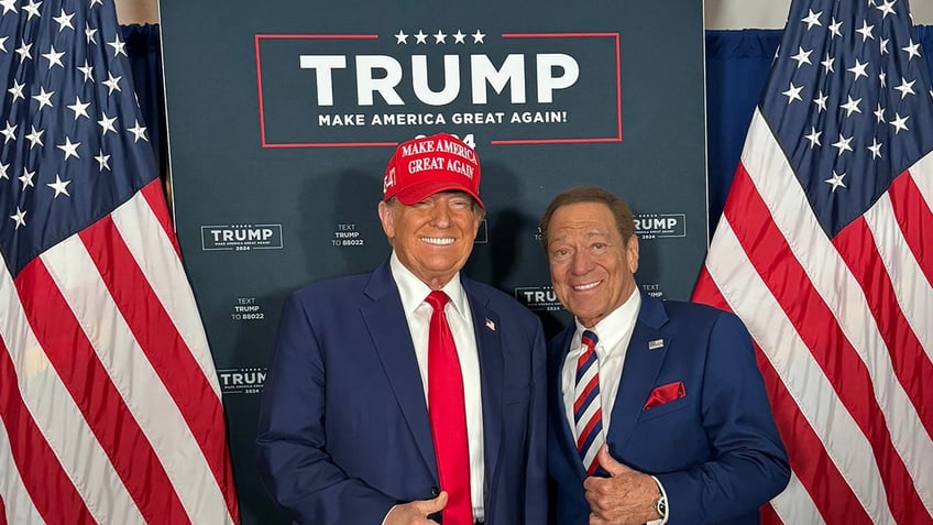 Donald Trump wearing a red Make America Great Again hat smiling and posing next to Joe Piscopo giving the thumbs up