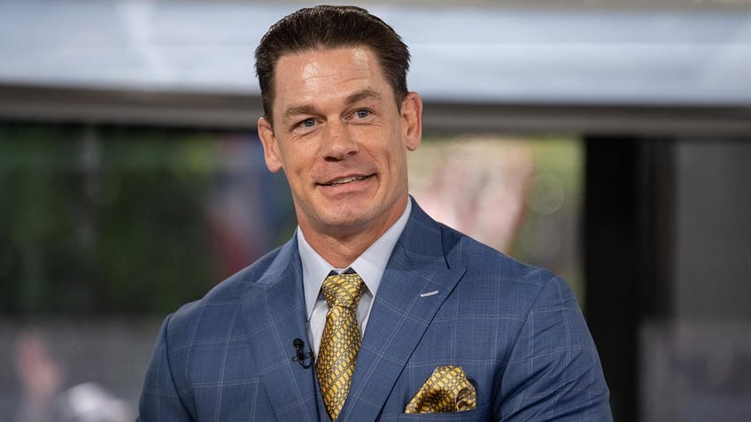 John Cena in a blue suit with a bright gold tie and pocket square smiles on set of the TODAY show