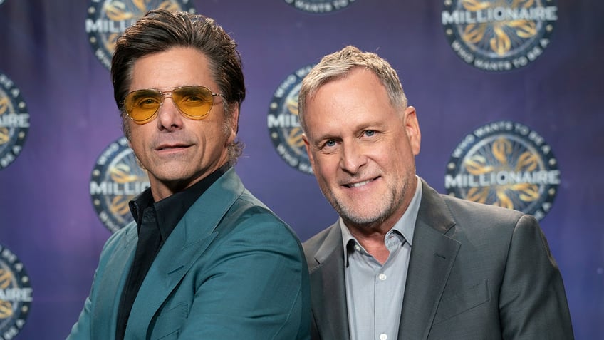 John Stamos in a turquoise jacket and yellow lensed sunglasses poses on the carpet with a smiling Dave Coulier in a grey a suit