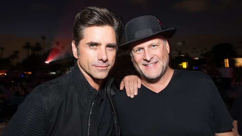 John Stamos in a black jacket puts his hand on Dave Coulier's shoulder, who is wearing a black shirt and hat