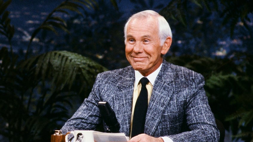 A close-up of Johnny Carson wearing a grey suit and sitting.