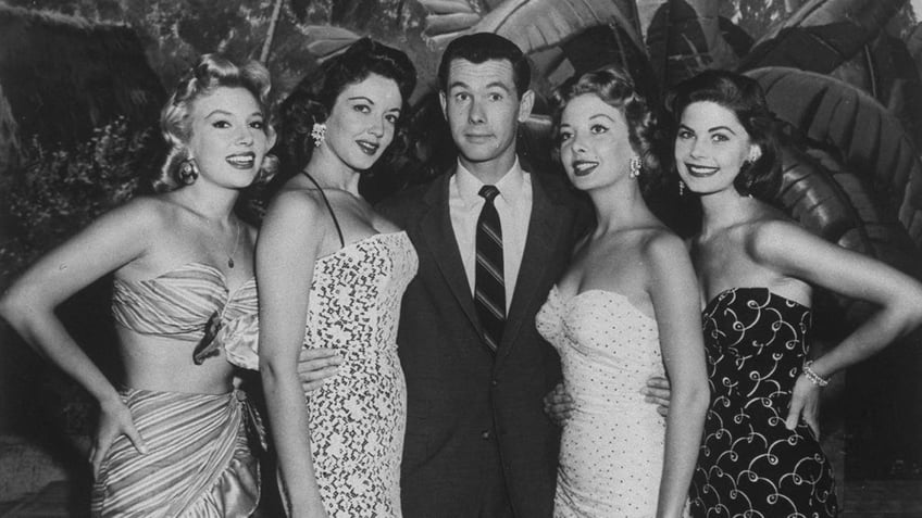 Johnny Carson in a suit surrounded by beautiful show girls.