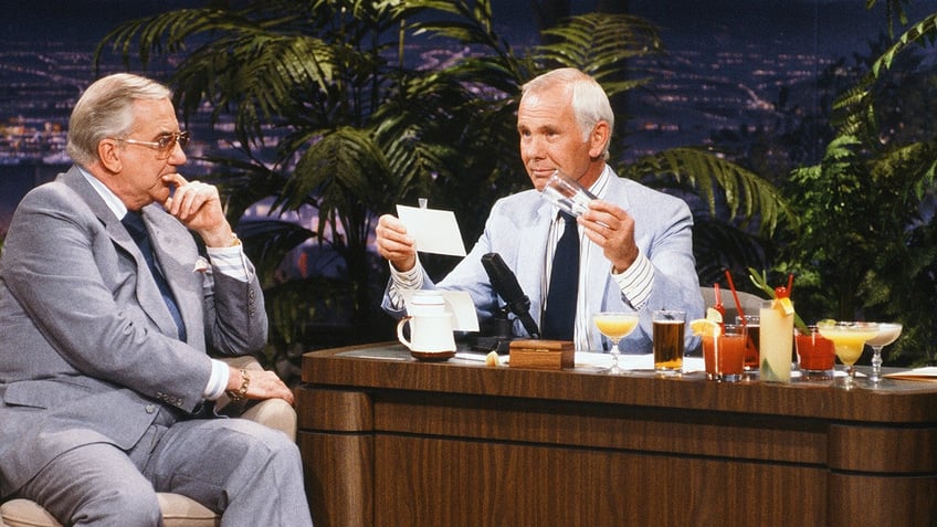 Johnny Carson sitting next to a row of drinks speaking with a guest.