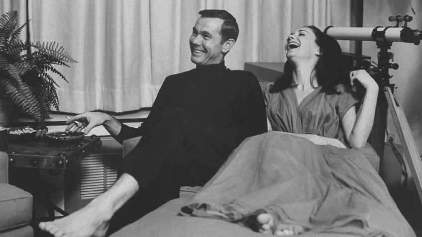 Johnny Carson and Joanne Copeland sitting in bed laughing barefoot