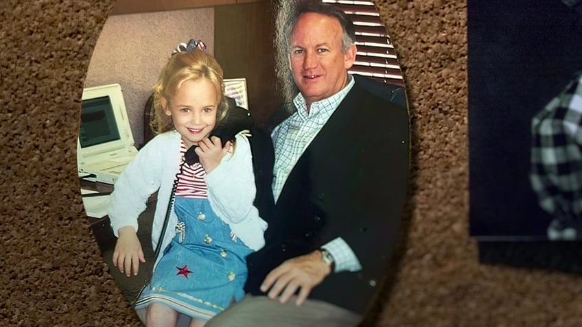 A photograph of JonBenet Ramsey holding a phone and sitting on her fathers lap.