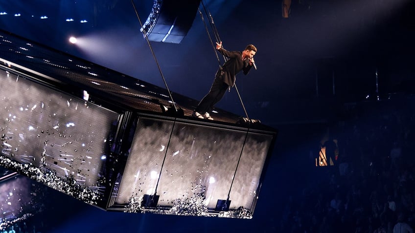 Justin Timberlake in a black suit and pants stands on a tilted stage above his concert crowd, secured to a harness