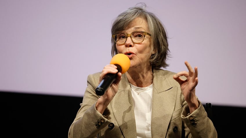 Sally Field in a tan jacket speaks into the microphone on stage and puts her fingers together