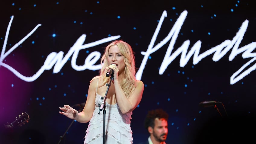 Kate Hudson in a light blue dress sings on stage and holds the microphone