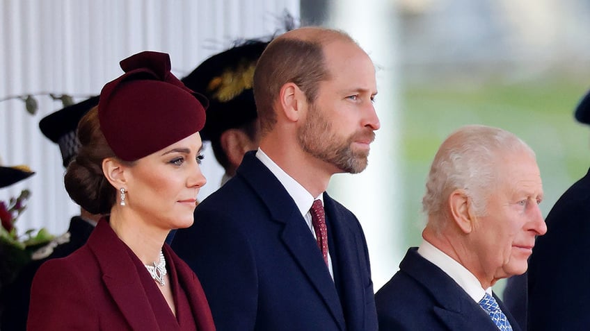 Kate Middleton wearing a burgundy coatdress with a matching hat standing next to Prince William and King Charles in suits.