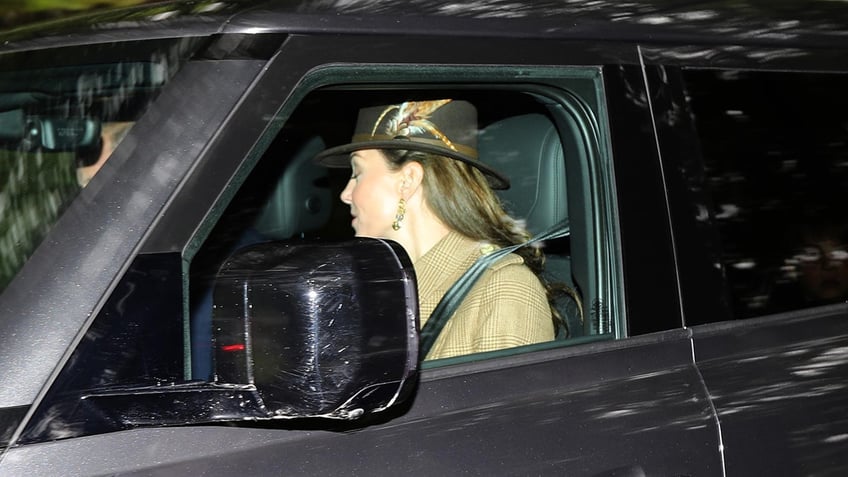 Kate Middleton looking away inside a car wearing a fedora and a beige jacket.