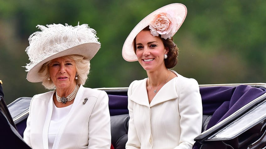 Camilla and Kate Middleton sitting together in a royal carriage wearing white dresses with giant fascinators to match.