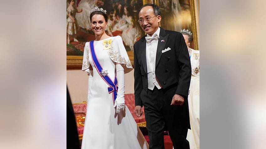 Princess Catherine walks in a long white dress with a blue sash and yellow brooch next to a man in black suit