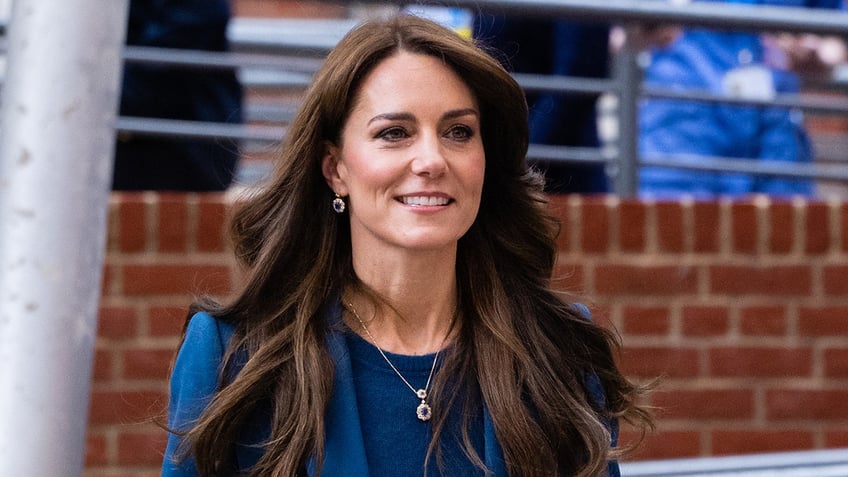 Kate Middleton in a royal blue shirt and matching jacket walks and partially smiles