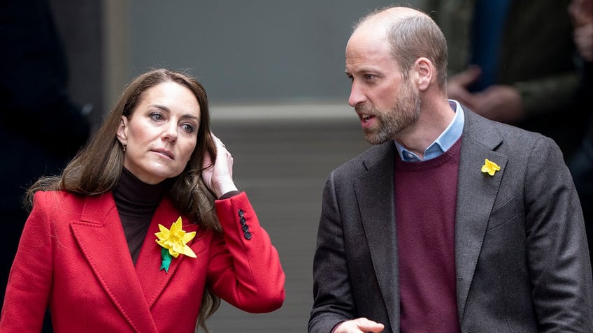 Prince William in a brown blazer, burgundy sweater and blue shirt, talking to Kate Middleton who is wearing a red coat and stroking her hair.