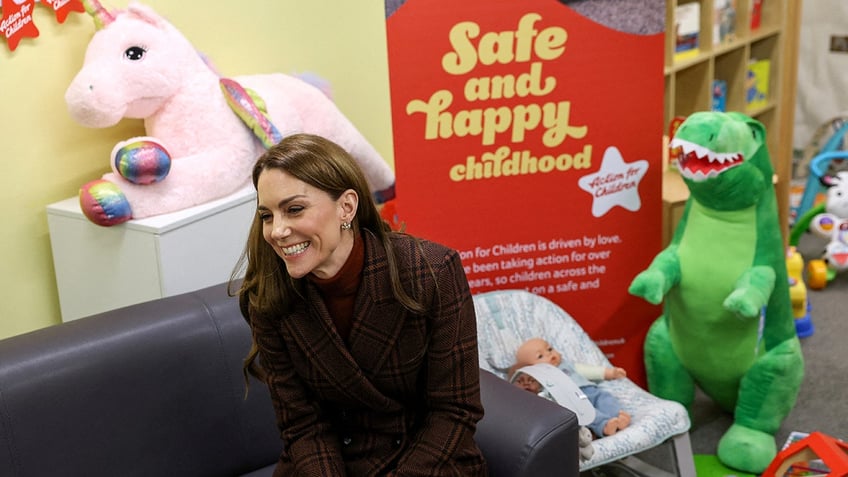 Kate Middleton wearing a brown coat dress smiling and sitting down next to toys.