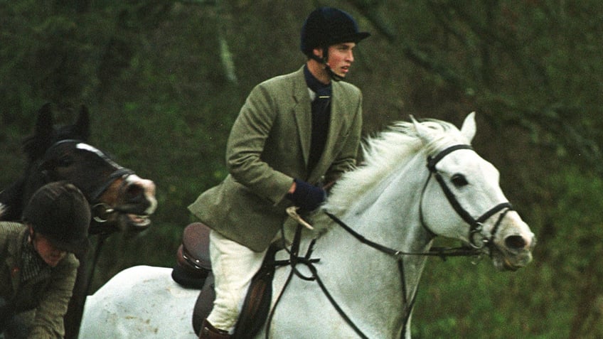 Prince William wearing a green blazer and white pants riding on a white horse while hunting.