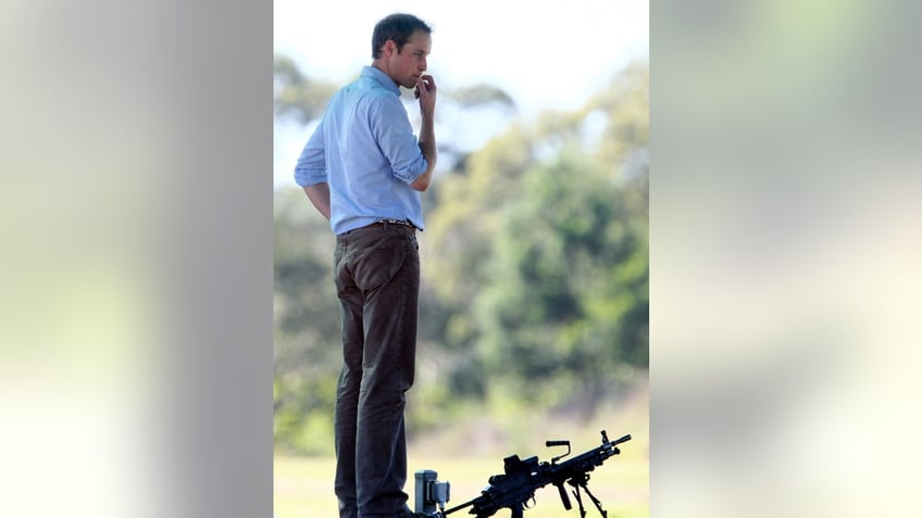 Prince William standing up outdoors wearing a blue shirt and grey pants next to a gun.