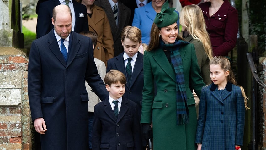 Prince William wearing a blue coat and Kate Middleton wearing a green coat with a matching scarf and hat walking outdoors with their three children.