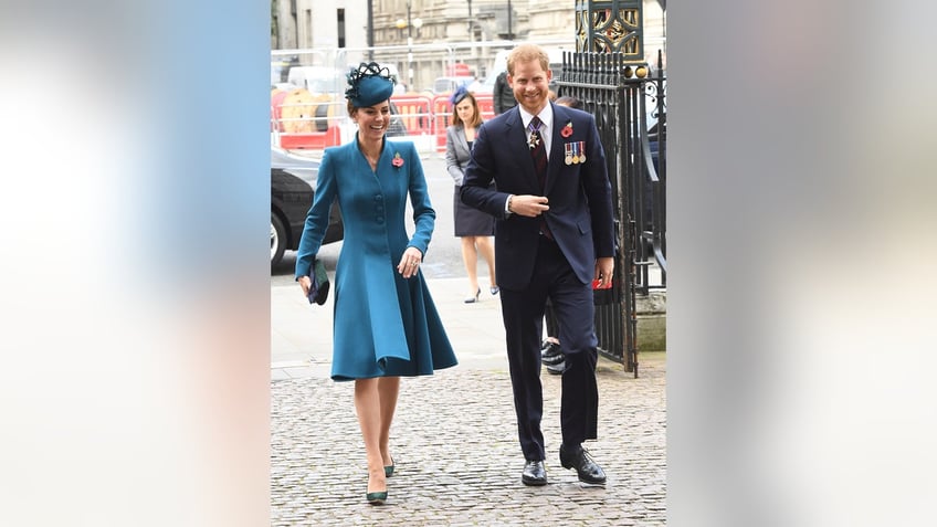 Kate Middleton in a blue dress and matching hat walking next to a smiling Prince Harry in a suit.