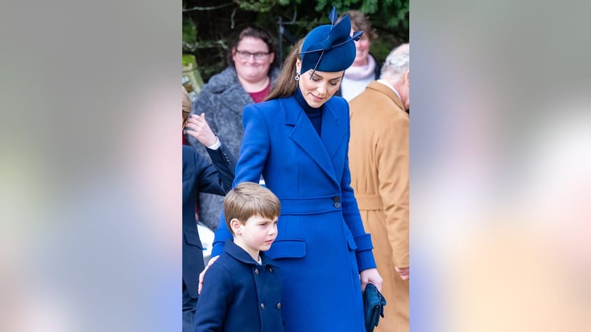 Kate Middleton in a blue coat dress touching Prince Louis back