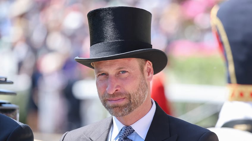 Prince William in a grey suit and black top hat sitting on a royal carriage.