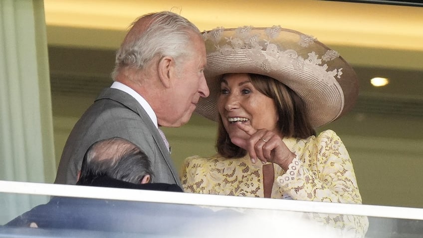 Carole Middleton in an ivory lace dress and matching hat smiling and chatting with King Charles wearing a grey suit.