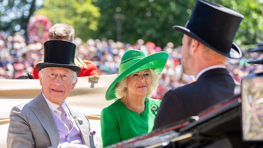 King Charles and Queen Camilla wearing formal suits and festive hats sitting across from Prince William on a royal carriage.