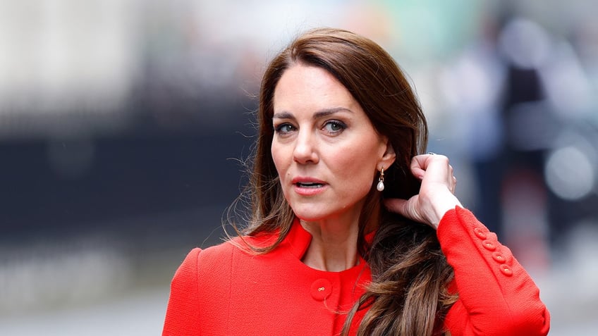 A close-up of Kate Middleton wearing a red suit stroking her hair.