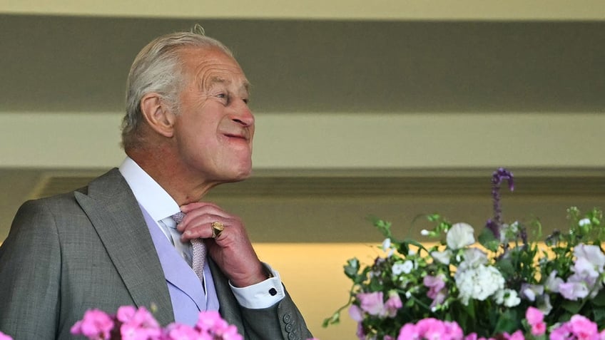 King Charles adjusting his tie in front of flowers.