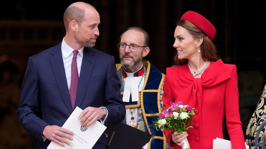 Kate Middleton in a red dress and matching hat holding a bouquet looking at Prince William in a dark suit.