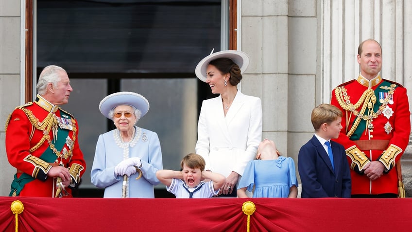 Kate Middleton chatting with King Charles at the Buckingham Palace balcony with Queen Elizabeth II and the royal children making funny facial expressions. Prince William, in uniform, looks on.