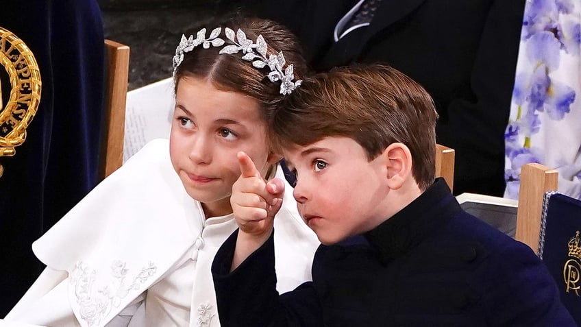 Prince Louis in a dark suit point up next to his sister Princess Charlotte who is wearing a white coat and a sparkling tiara.