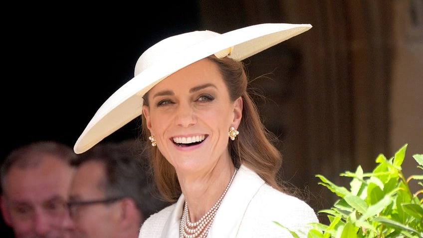 A close-up of Kate Middleton smiling wearing a white coat dress and a matching hat.