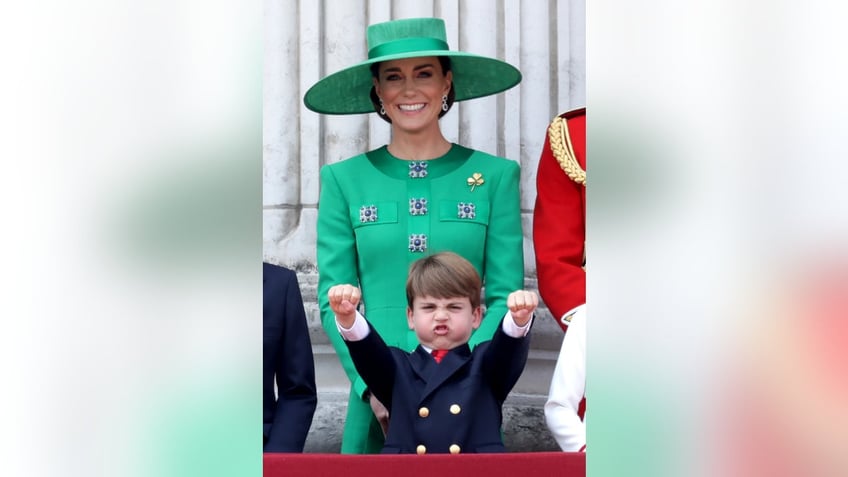Kate Middleton wearing a green dress with a matching hat smiling as Prince Louis shows off his fists on the palace balcony