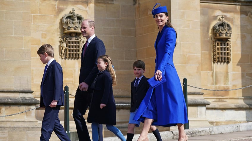 Kate Middleton in a cobalt blue coat dress walking with her husband Prince William and their three children