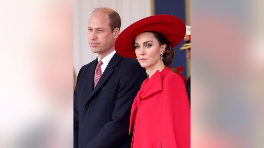 Kate Middleton wearing a red dress and matching hat looking serious next to Prince William in a dark suit and red tie