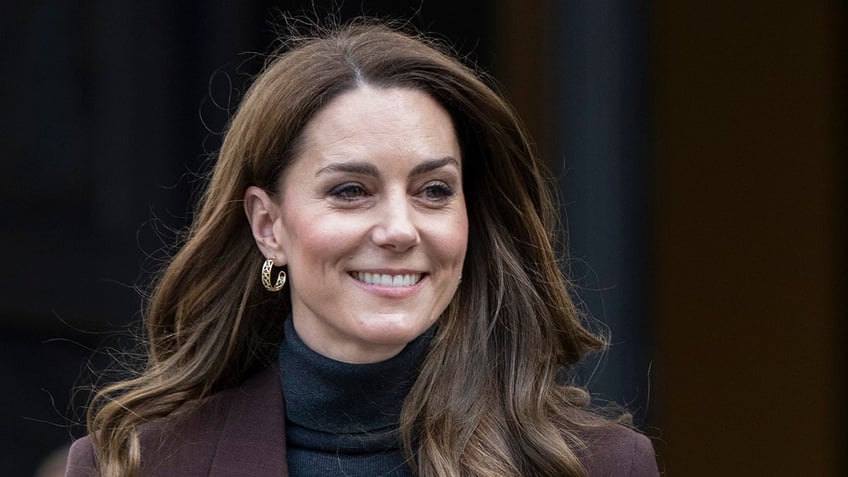 A close-up of Kate Middleton smiling wearing a black turtleneck and a brown blazer