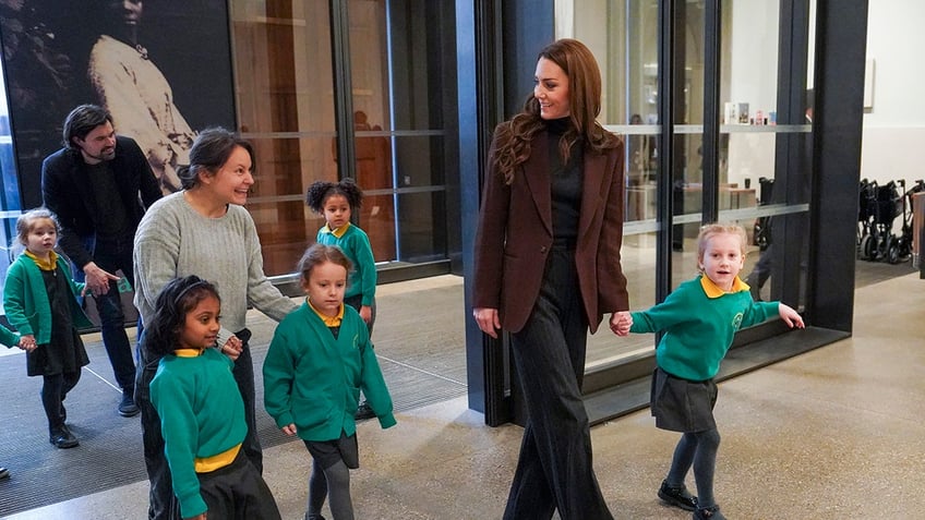 Kate Middleton wearing a brown blazer a black shirt and black pants holding a little girl's hand