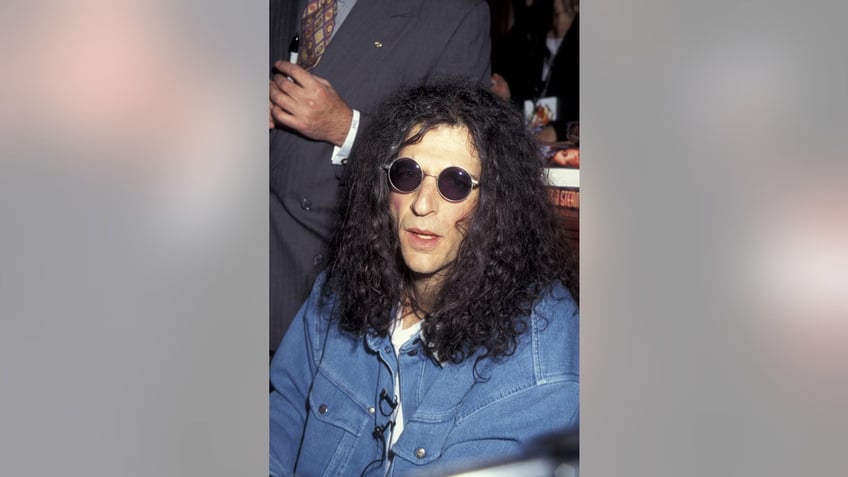 Howard Stern wearing a blue denim shirt and round sunglasses