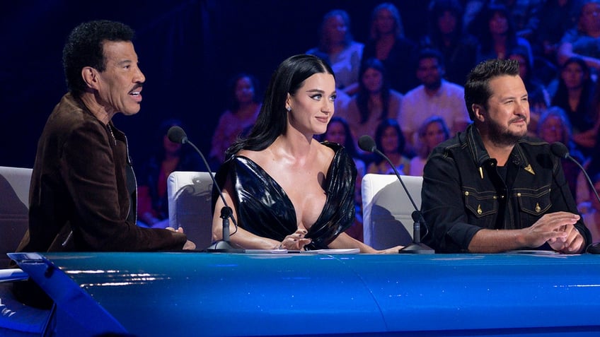 Lionel Richie in a brown suede jacket sits behind the judge's desk with Katy Perry in a black dress and Luke Perry in a dark jean jacket