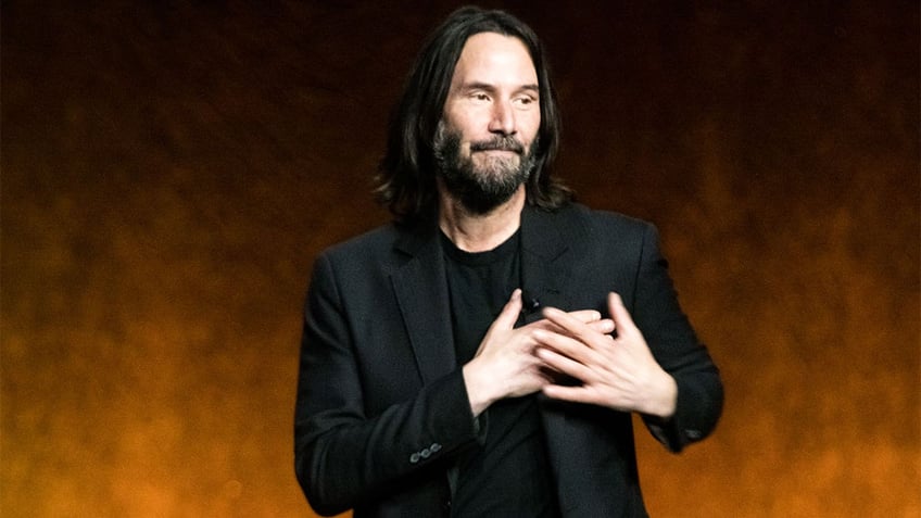 Keanu Reeves in a black blazer and t-shirt puts his hands to his chest (near his heart) on stage