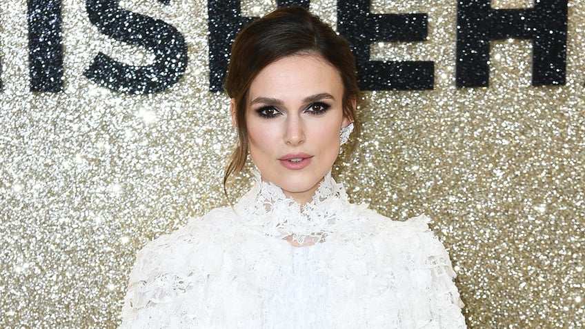 Keira Knightley in a lace white dress looks directly at the camera on the red carpet