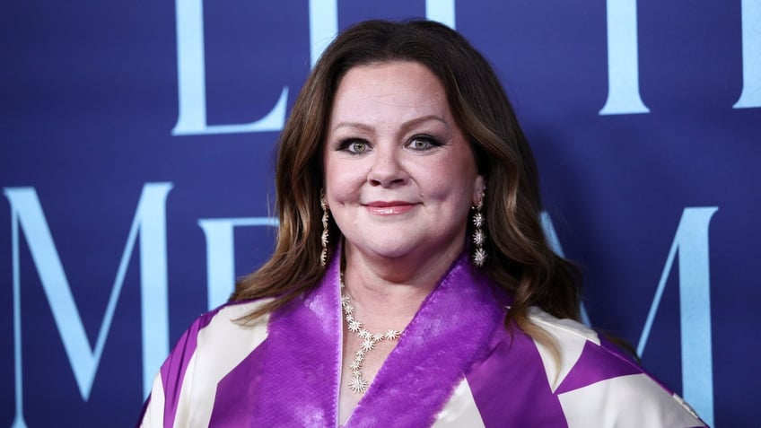 Melissa McCarthy in a purple patterned dress soft smiles on the carpetMelissa McCarthy in a purple patterned dress soft smiles on the carpet