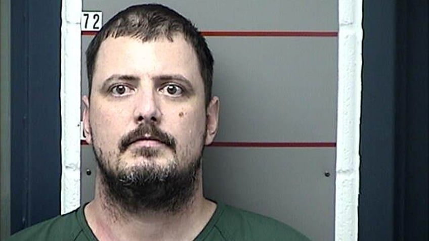 Jesse Kipf stares blankly with a beard and mustache as he stands against the wall in a mugshot, wearing a green jail uniform