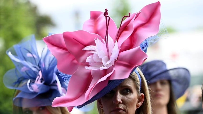 Two women wear large flower-like hats at the Kentucky Derby. One woman wears a pink hat while another woman wears a purple hat.