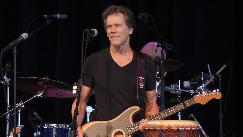 Kevin Bacon holds a guitar while performing on stage.