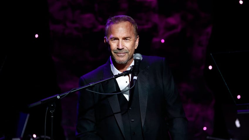 Kevin Costner speaks on stage wearing back tuxedo