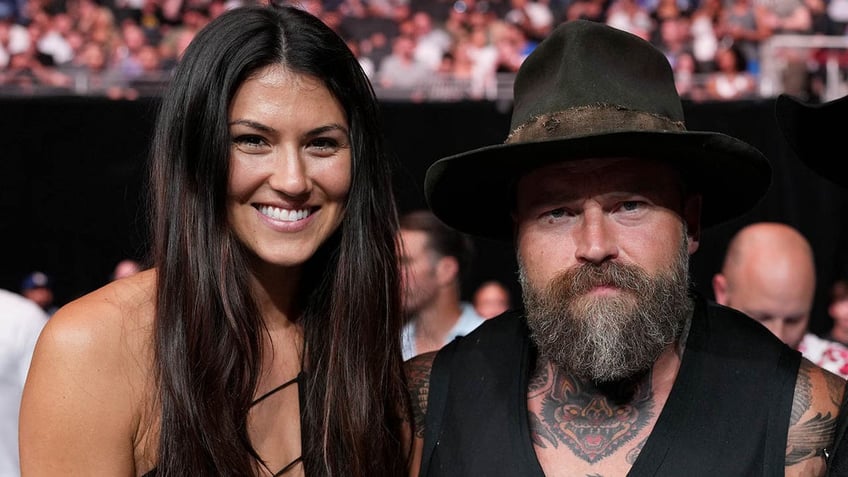 Kelly Yazdi smiles next to husband Zac Brown in a black vest and cowboy hat