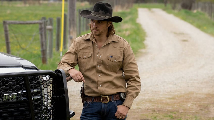 Luke Grimes in a black cowboy hat and tan shirt leans against a car in a scene from "Yellowstone"