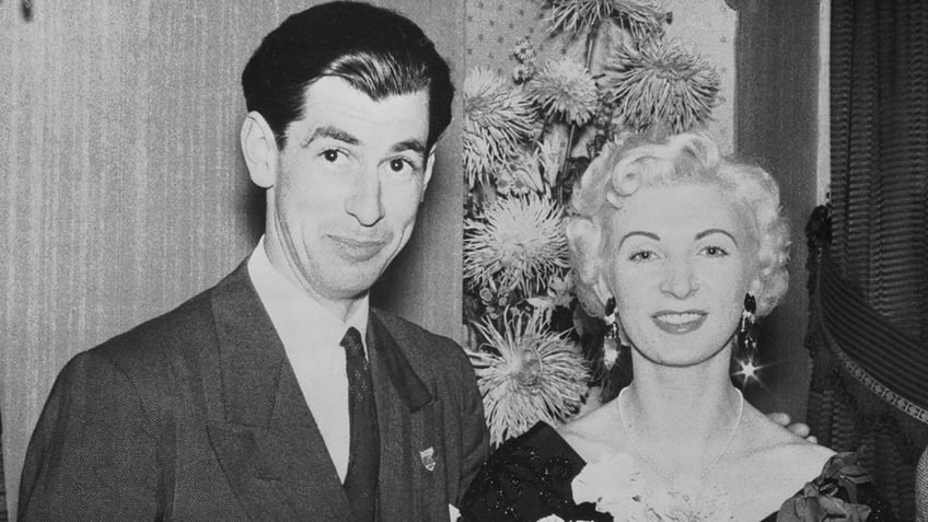 David Blakely in a suit posing next to a smiling Ruth Ellis and a floral arrangement.